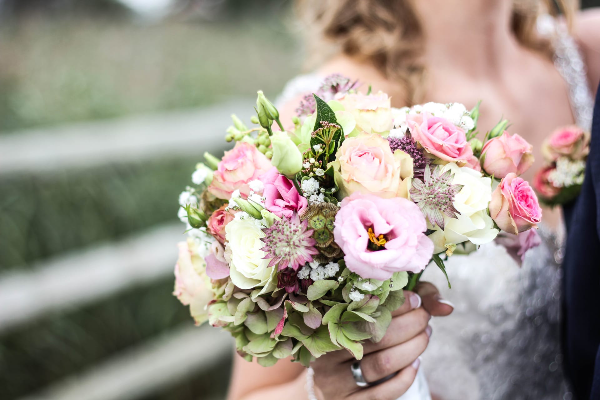 Un bouquet de mariée de roses pâles, d’hortensias et de chardons tenu à deux mains devant une robe blanche