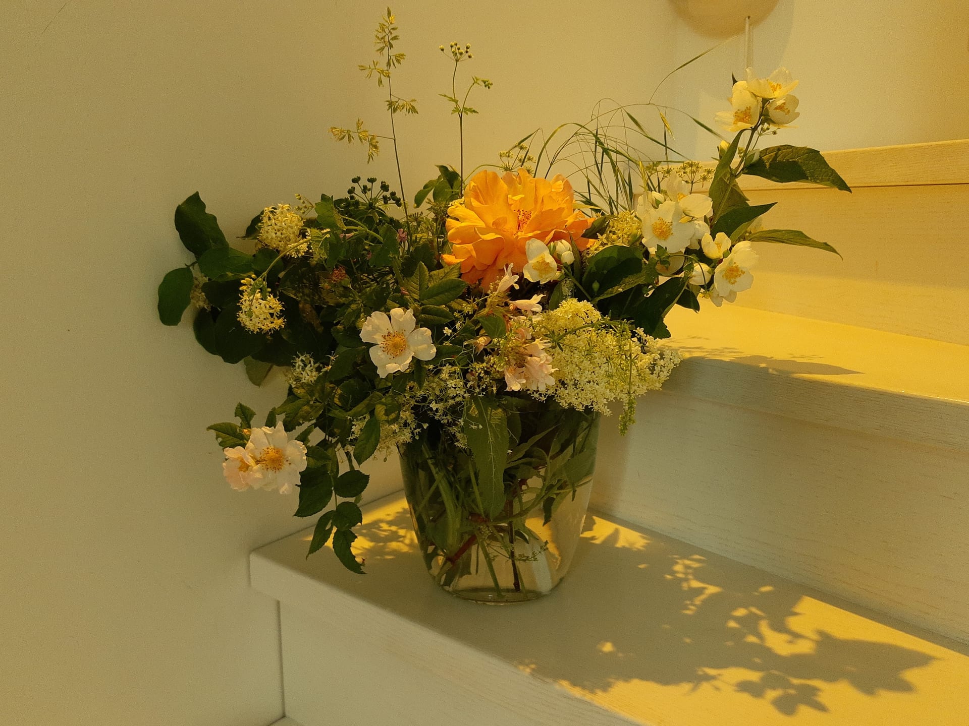Un bouquet de fleurs de saison orange, jaunes et blanches dans un vase en verre posé sur une marche ensoleillée
