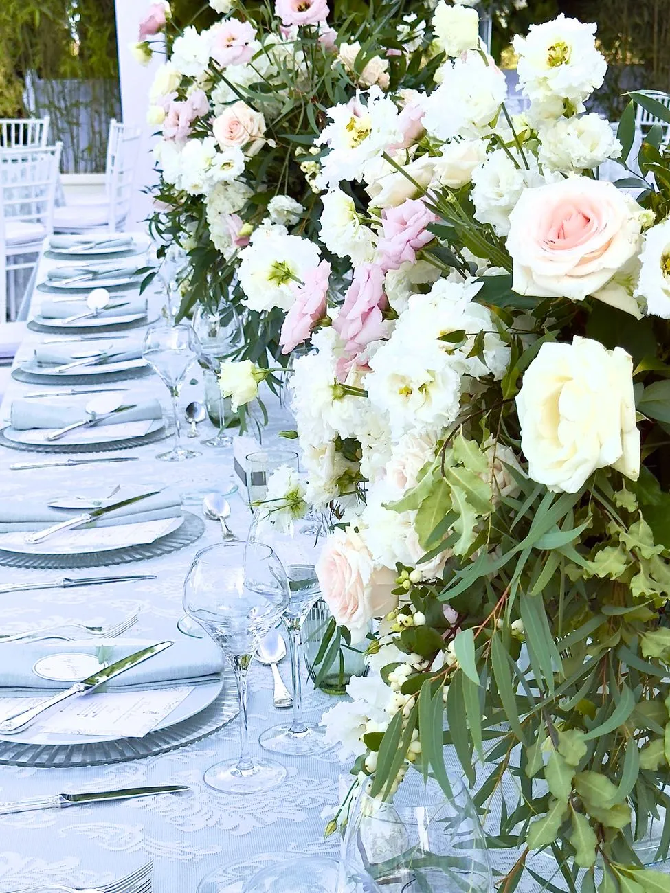 Longue table de mariage dressée avec des assiettes alignées et un haut centre de table fleuri blanc et rose