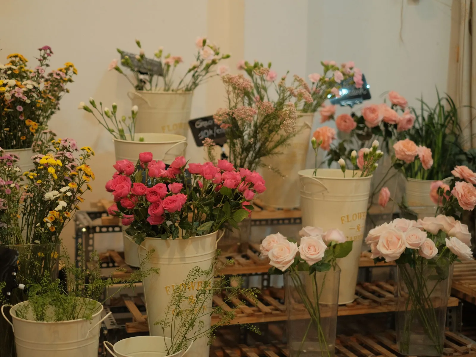 Œillets roses et rouges, roses pâles et fleurs des champs dans des seaux crème posés sur des étagères en bois